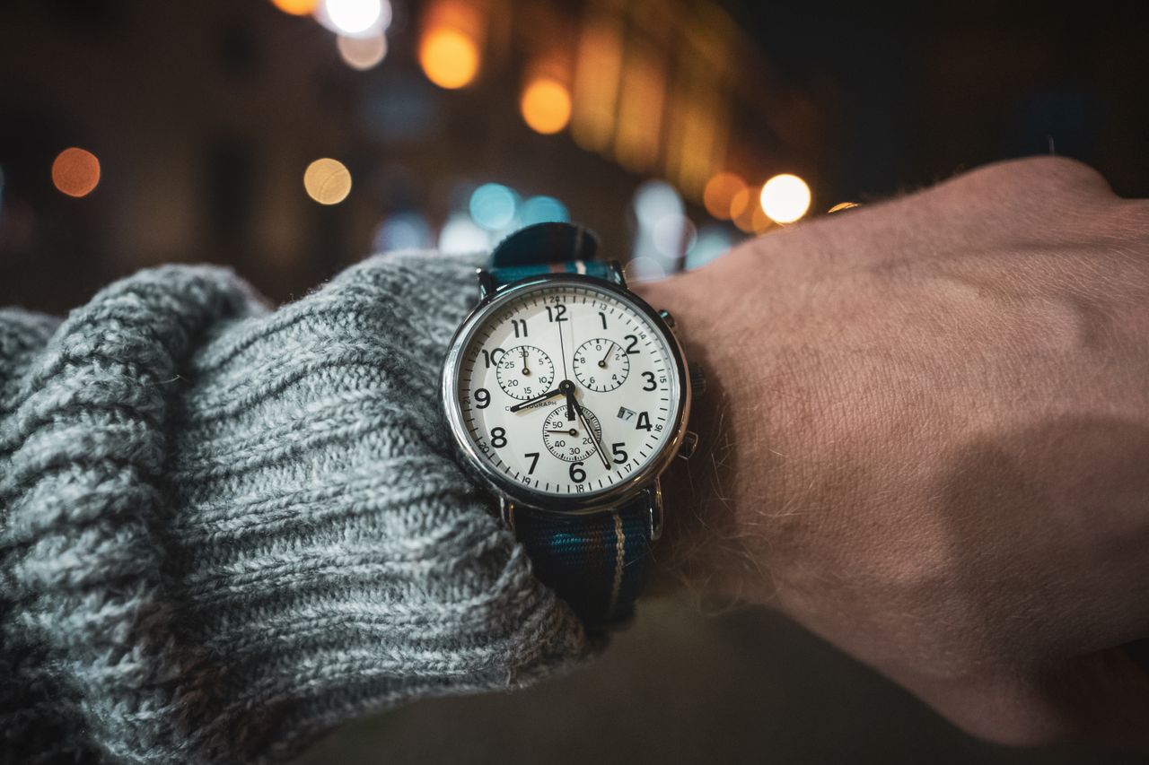 who-we-are A detailed close-up of an analog wristwatch on a wrist in a night setting with bokeh background.